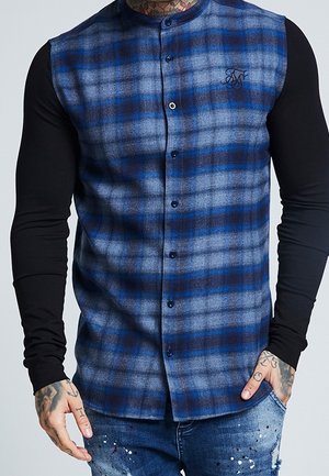 Homme portant un gilet sans manches à carreaux bleu et noir boutonné sur une chemise noire à manches longues, avec des mains tatouées et un jean éclaboussé.