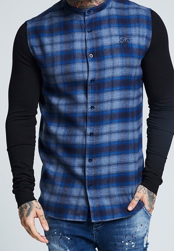 Homme portant un gilet sans manches à carreaux bleu et noir boutonné sur une chemise noire à manches longues, avec des mains tatouées et un jean éclaboussé.