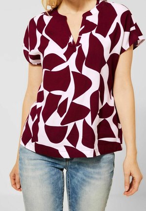 Kvinde iført kortærmet bluse med bordeaux og hvidt abstrakt mønster kombineret med lyseblå jeans mod ensfarvet baggrund.