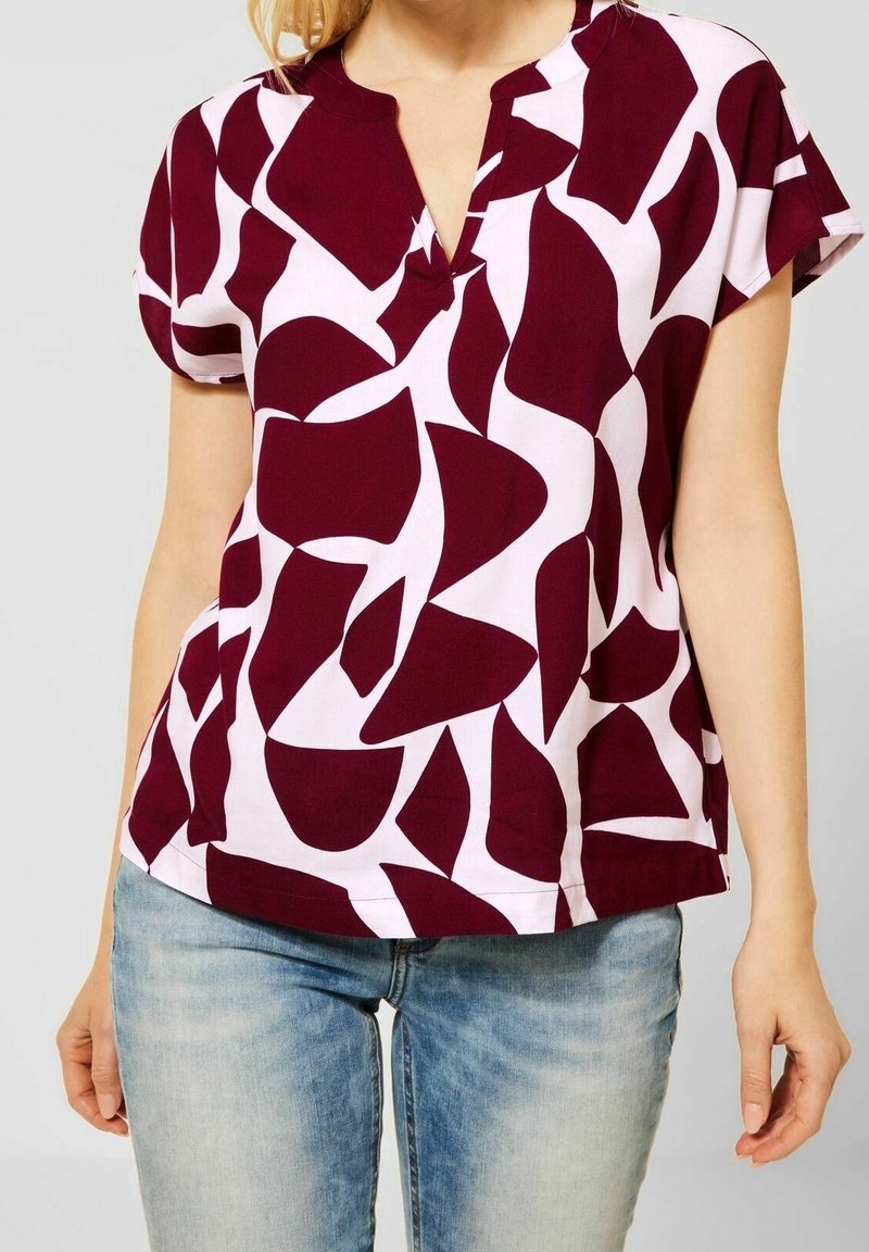 Femme portant un chemisier à manches courtes avec un motif abstrait bordeaux et blanc assorti à un jean bleu clair sur un fond uni.