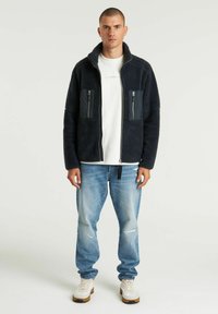 Giacca in pile blu navy con colletto alto, due tasche sul petto con zip e texture morbida, abbinata a jeans chiari strappati e sneakers bianche.