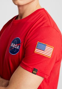 Detailní záběr osoby v červeném tričku NASA s logem NASA na hrudi a výšivkou americké vlajky na rukávu.