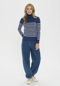 Pulover cu guler înalt cu dungi navy, realizat dintr-un material tricotat, asortat cu pantaloni din denim albastru, largi și adidași de culoare crem.