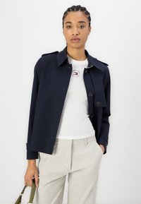 Marineblå trenchcoat med en avslappet passform, knappelukking og to frontlommer, kombinert med en hvit t-skjorte med grafikk og lyse beige bukser.
