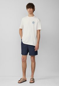 Witte katoenen T-shirt met een kleurraf rond grafisch ontwerp, gecombineerd met donkerblauwe korte broek en bruine sandalen. Casual zomeroutfit ontwerp.