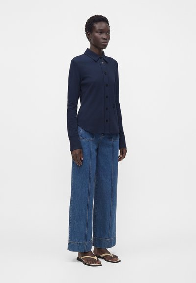 Femme debout portant une chemise bleu marine boutonnée à manches longues, un jean bleu à jambes larges et des sandales beige tongs sur un fond uni.