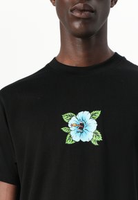 Sort bomulds t-shirt med et blåt blomstermotiv, der har en mund og gule detaljer, omgivet af grønne blade; rund halsudskæring.