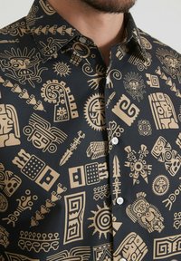 Chemise à manches courtes noire avec des motifs tribaux dorés. Fabriquée en tissu lisse, avec un boutonnage à l'avant et un col pointe.