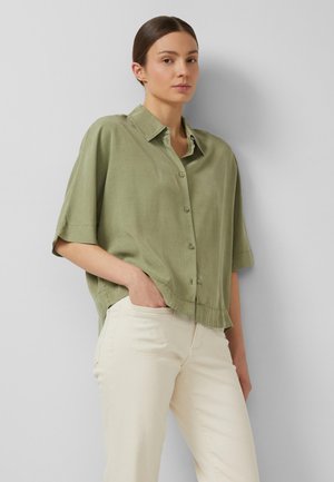 Vrouw met donker haar in een lage knot, draagt een losse olijfgroene blouse met knopen en crèmekleurige broek, staand met één hand in de zak.