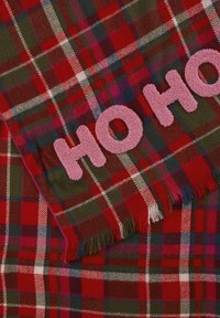 Écharpe à carreaux rouges et verts avec le texte rose "HO HO". Le matériau est doux avec des bords à franges, affichant un motif tartan classique.