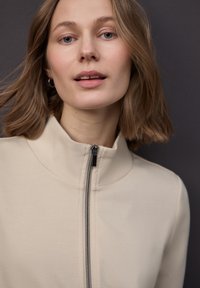 Giacca beige con zip e collo alto, realizzata in tessuto liscio con dettaglio della zip nera a contrasto. Primo piano del viso e dei capelli.