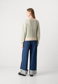 Suéter creme tricotado com padrão zigzag verde, bainha e punhos canelados. Combinado com jeans largos azuis com bainhas cinzentas desfiadas e sapatos pretos.