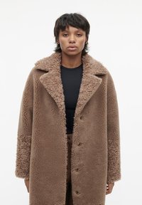 Bruine sherpa jas met klepcol, knoopsluiting en zachte, donzige textuur; gedragen over een zwarte top. Eenvoudige, relaxte pasvorm.