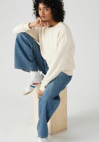 Crème kleurige sweatshirt met geborduurd logo, gecombineerd met lichtblauwe wijde jeans en witte sneakers met rode en blauwe strepen.