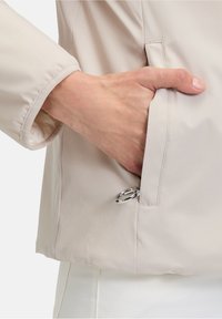 Veste beige clair avec une texture douce, dotée d'une poche latérale et d'un accent en anneau argenté à l'ouverture. Poignets de manches élastiqués.