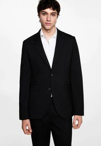 Sacou negru cu rever înclinat, două butoane și un buzunar pe piept. Purtați peste o cămașă albă cu guler, cu un croi ajustat.