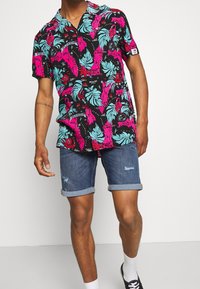 Kurzarmhemd mit schwarzem Grund und lebendigen pinken sowie blauen tropischen Blatt- und Flamingomustern; kombiniert mit Jeansshorts.