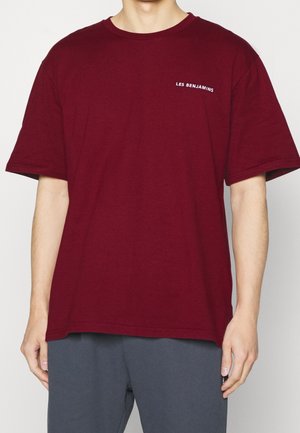 Mann trägt ein schlichtes burgunderfarbenes T-Shirt mit kurzem Ärmel und dem Schriftzug "LES BENJAMINS" auf der oberen linken Brustseite sowie graue Hosen.