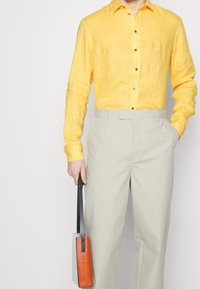 Žlutá lněná košile na knoflíky s texturovaným povrchem, kombinovaná s béžovými kalhotami. Drží malou dvoubarevnou tašku s oranžovým akcentem.