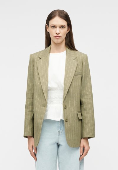 Femme portant un blazer beige à rayures fines sur un haut blanc et un jean bleu clair, debout devant un fond clair uni.