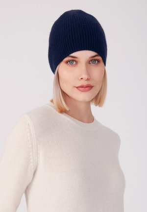 Beanie a coste di un blu scuro, realizzato in materiale morbido, indossato aderente, abbinato a un maglione di un crema chiaro, che presenta un design semplice e texture.
