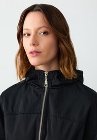 Zwarte hoodie-jack met een ritssluiting aan de voorkant en gouden hardware. Gemaakt van een gladde stof, heeft het een gestructureerd ontwerp en een relaxte pasvorm.