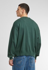 Sudadera verde de cuello redondo en un corte relajado, confeccionada en tejido suave. Presenta puños y dobladillo acanalados, con un emblema en los bolsillos traseros de los vaqueros.