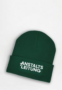 Green knit beanie with a ribbed texture and a folded cuff. Features a rectangular patch with "ANSTALTSLEITUNG" in bold white lettering.