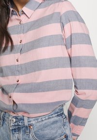 Femme portant une chemise boutonnée à rayures horizontales rose et grise, rentrée dans un jean en denim bleu clair.