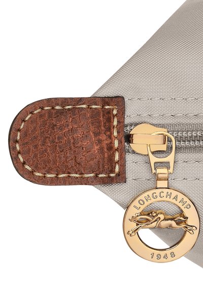 Hellgraue Stofftasche mit braunem, strukturiertem Lederakzent, goldfarbener Reißverschluss-Ausstattung und einem runden goldfarbenen Anhänger mit geprägt eingraviertem Logo und Datum.