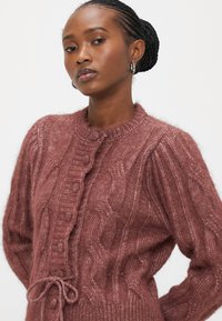 Πλεκτή ζακέτα σε απαλό βυσσινί χρώμα, με ανάγλυφο σχέδιο καλωδίων και στρογγυλή λαιμόκοψη, κλείνει με κουμπιά και κορδέλα στη μέση.