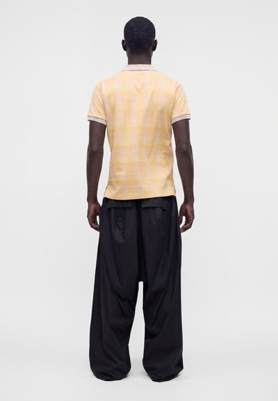 Homme vu de dos portant une chemise à carreaux jaune à manches courtes et un pantalon large noir ample sur un fond clair uni.