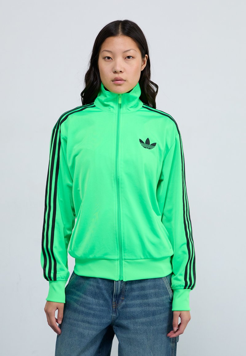Grön zip-up jacka i slätt material, med svarta ränder längs ärmarna och en liten svart logotyp på bröstet.