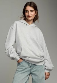 Felpa grigia chiara realizzata in tessuto morbido, con un ampio cappuccio, tasca a marsupio e polsini a costine. Abbinata a jeans in denim blu chiaro.