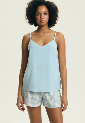 Kobieta o kręconych włosach, ubrana w jasnoniebieską koszulkę na ramiączkach i niebiesko-białe szorty w liściasty wzór, stoi na jednolitym tle.