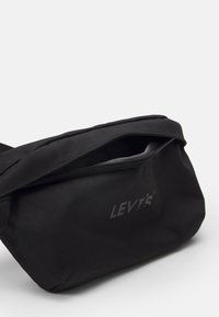 Mustat vyölaukku, joka on valmistettu tekstuurikankaasta, varustettu päällimmäisellä vetoketjulla ja näkyvällä logolla. Kompakti muotoilu, jossa on säädettävä hihna.