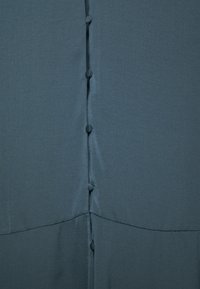 Gros plan sur un tissu bleu sarcelle foncé avec une ligne verticale de boutons recouverts et une couture horizontale près du bas.