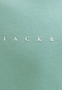 Λευκά γράμματα που σχηματίζουν τη λέξη "JACK &" σε φόντο υφασμάτινου φόντου σε ανοιχτό τιρκουάζ με υφή, τοποθετημένα σε ευθεία οριζόντια γραμμή.