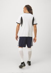 Camisola atlética branca com painéis laterais pretos, calções azul-marinho, meias brancas até ao joelho e chuteiras pretas. Foco na funcionalidade desportiva.