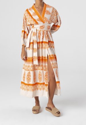 Vestito lungo avvolgente in tessuto crema con motivi floreali arancioni e righe. Presenta maniche lunghe, uno spacco laterale e una vita con cintura. Abbinato a delle ciabatte beige.