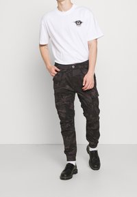 Valkoinen lyhythihainen t-paita, jossa rintakehällä graafinen logo, yhdistettynä mustiin maastokuvioisiin cargo-housuihin, joissa on taskut ja joustavat resorit. Mustat kengät.