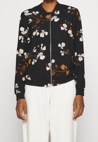 Chaqueta bomber negra con patrones florales en blanco y marrón, cierre de cremallera en la parte delantera, puños y dobladillo acanalados. Fabricada con un tejido suave.