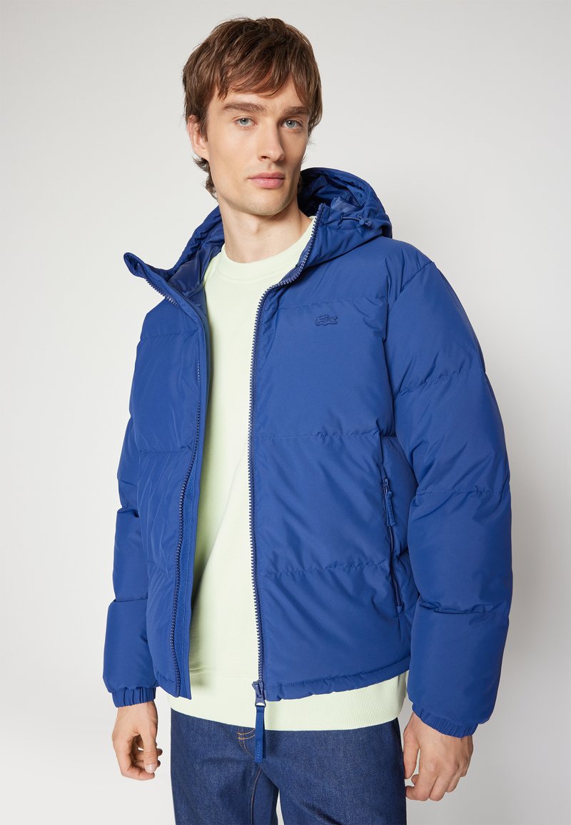 Chaqueta de plumas azul real con capucha, con cierre de cremallera en la parte delantera, puños elásticos y detalle de logo. Lleva encima un sudadera verde claro.