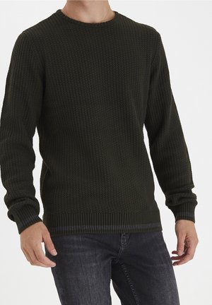 Mężczyzna w ciemnozielonym, teksturowanym swetrze z dzianiny i ciemnych dżinsach, stojący na tle jednolitej białej ściany.