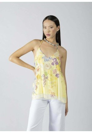 Donna con lunghi capelli neri che indossa una canottiera gialla a fiori e pantaloni bianchi, in posa con la mano sul fianco davanti a uno sfondo semplice.