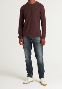 Chemise à manches longues bordeaux en tissu texturé, col rond, avec un patch logo noir, assortie à un jean bleu et des baskets blanches.