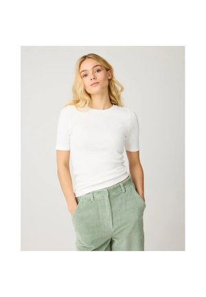 Jeune femme aux longs cheveux blonds portant un haut blanc à manches courtes et un pantalon en velours côtelé vert clair, debout les mains dans les poches.