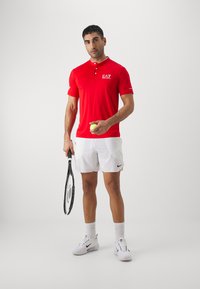Maglietta polo rossa con colletto e tre bottoni, abbinata a pantaloni corti bianchi. Tiene in mano una palla da tennis e una racchetta, indossando scarpe e calze bianche Nike.
