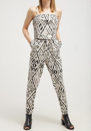 Kobieta ubrana w czarnobiały, geometryczny, bezrękawnikowy kombinezon z sznurkiem w talii i czarne botki na odkryte palce, ręce w kieszeniach.