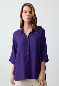 Chemise violette à boutons avec un col, coupe ample et manches trois-quarts, fabriquée à partir d'un tissu doux et lisse.